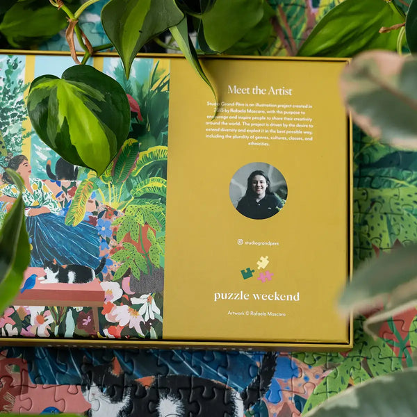 Jigsaw puzzle box with 'Meet the Artist' section and colorful illustration on a completed portion of the puzzle among a green leafy background. Photography by Esther Louise