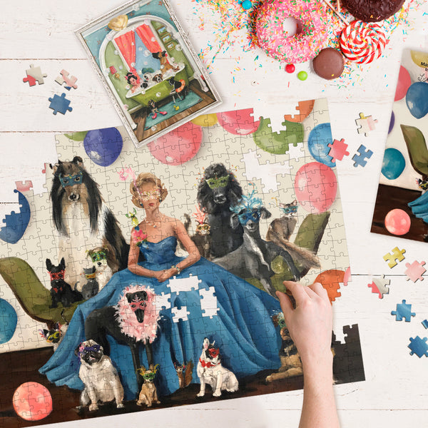 Hand putting together bright jigsaw puzzle showing artwork of a woman in party dress next to a group of dogs and cats. Puzzle pieces scattered around and next to plate of donuts.