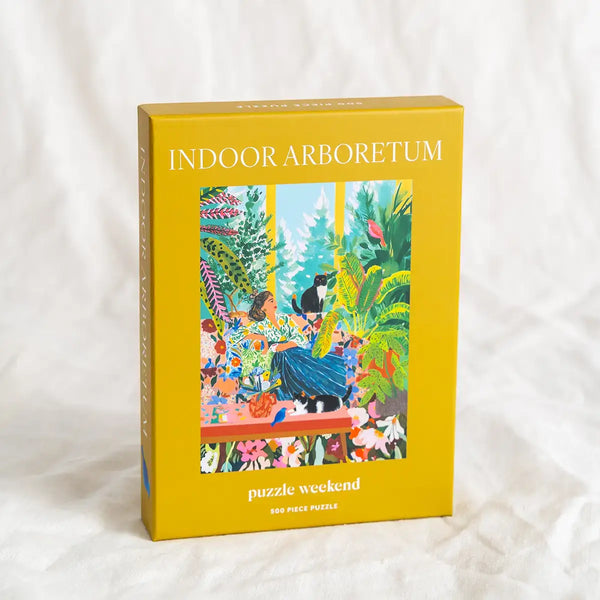 Yellow puzzle box titled 'Indoor Arboretum' with a colorful illustration of a woman resting on a sofa surrounded by plants on a white background. Photography by Esther Louise