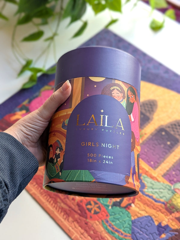 Hand holding a Laila Luxury puzzle box for the puzzle 'Girls Night' shown over portion of the completed puzzle on a white desk with plants.