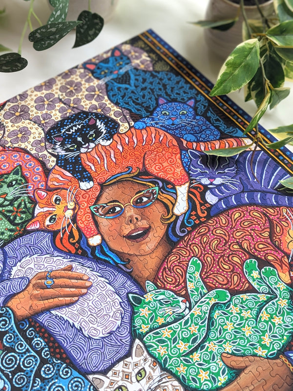Colorful jigsaw puzzle artwork of a woman with cats and abstract patterns on a desk surrounded by plants
