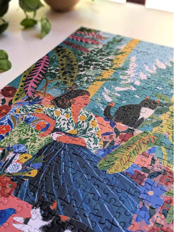A colorful completed jigsaw puzzle featuring a stylized illustration of a woman in a floral blouse and dark skirt, surrounded by tropical leaves and cats, photographed on a light tabletop with a plant leaf in the corner.