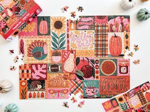 A colorful autumn-themed jigsaw puzzle with pieces scattered around it on a white surface, alongside the puzzle box and partial matching puzzle pieces. The puzzle features a patchwork design of pumpkins, leaves, plaid patterns, and fall motifs in orange, pink, teal, brown, and cream tones.
