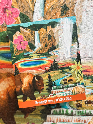 A 1000-piece jigsaw puzzle by Spaghetti Tree Puzzles, featuring a colorful landscape illustration with a bison, waterfall, hot spring, and mountains, displayed over a partial view of the completed puzzle.