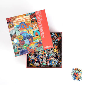 An open puzzle box showing the pieces being held in the bottom of the box and the lid of the box resting on top showing a colorful collage of national park themed stickers. A small group of puzzle pieces are set to the bottom right. 