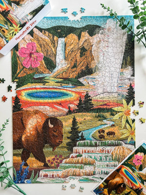 A completed multicolor jigsaw puzzle showing a scenic landscape with a bison, waterfall, rainbow, and terraced pools, styled on a white surface with puzzle pieces, plants and the puzzle poster around it.