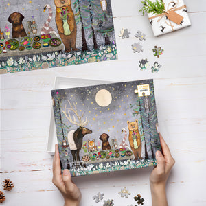 Top view of hands holding Moonlit Kingdom puzzle box lid on top of white wood desktop with puzzle pieces scattered around. 