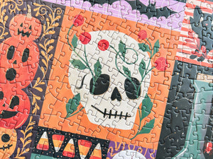 A close-up of a completed jigsaw puzzle featuring a skull with roses growing out the eye holes on an orange background, surrounded by Halloween-themed illustrations including a jack-o'-lantern and candy corn.