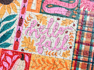 A close-up of a completed jigsaw puzzle featuring a colorful autumn design with the words 'Hello fall' in pink script, orange pumpkins, teal leaves, and plaid patterned sections.
