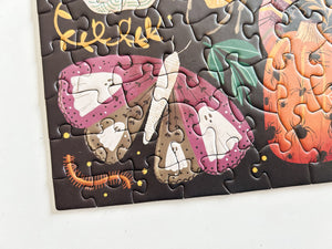 A close-up of a completed jigsaw puzzle showing a whimsical Halloween illustration with a purple-pink butterfly, white ghosts, an orange pumpkin, and a small orange centipede on a dark background.