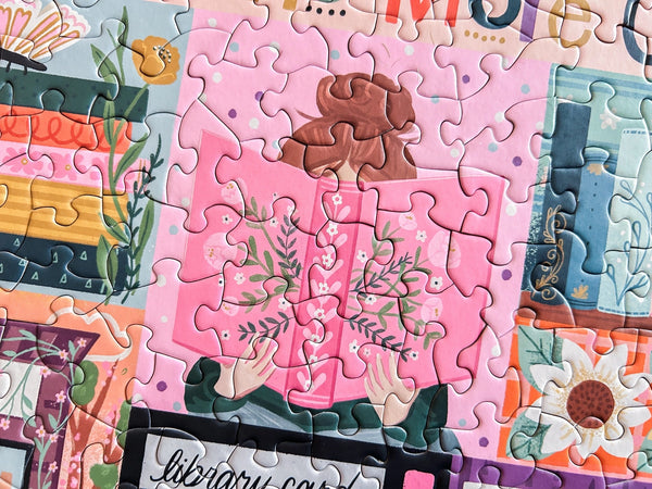 Jigsaw puzzle with a pink section featuring a person holding a book with a floral cover, surrounded by colorful puzzle pieces.
