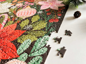 A close-up of a completed jigsaw puzzle showing a colorful floral and botanical design, with a few loose puzzle pieces on a white surface next to a pinecone and evergreen sprig.