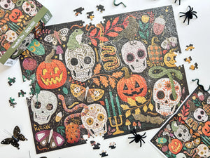A Halloween-themed jigsaw puzzle with colorful skull, pumpkin, snake, candle, and butterfly illustrations, shown partially assembled on a white surface with loose puzzle pieces and the puzzle box around it.
