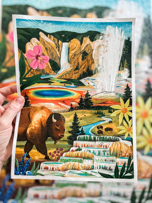 A hand holds a colorful illustrated art print depicting Yellowstone National Park, featuring a bison, waterfall, geothermal pool, flowers, and layered terraces, with a blurred multicolor background.