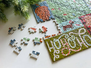A Christmas-themed jigsaw puzzle set on a white surface, with several pieces scattered nearby and evergreen branches in the top left corner. The puzzle features holiday illustrations and the words 'HO HO HO' in large white letters with red and green accents.