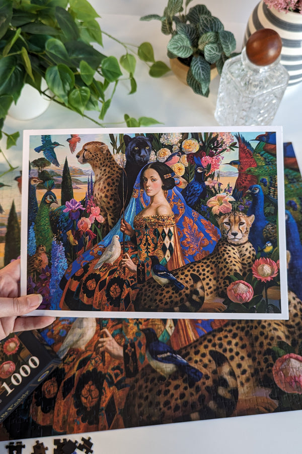 High-Res Art Print of the puzzle held in a hand over the puzzle. Print features a regal woman staring at the viewer surrounded by large cats and elegant birds.