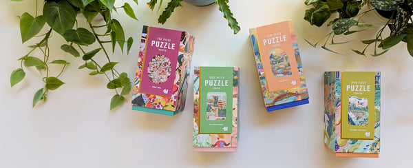 Four colorful Lantern Press puzzle boxes on a white surface with green plants in the background