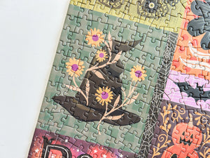A close-up of a completed jigsaw puzzle showing a Halloween-themed design with a witch's hat, yellow flowers, spiderwebs, bats, and a jack-o'-lantern and a multicolored background on a white surface.