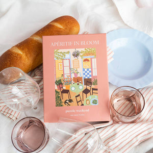 Puzzle box titled 'Apéritif in Bloom' with a scenic cafe illustration, surrounded by bread, glasses, and a plate on a white surface.
