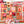 A hand holds an art print featuring a colorful autumn patchwork design with pumpkins, sunflowers, turkeys, leaves, and fall-themed illustrations. The background is a softly blurred mostly completed version of a puzzle featuring the same artwork.