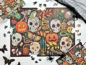A Halloween-themed jigsaw puzzle with scattered pieces on a white surface, alongside the puzzle box, a black spiderweb material, and decorative spider and butterfly props. The puzzle design features sugar skull, jack-o'-lantern, candelabra, snakes, and autumn leaves on a dark background.