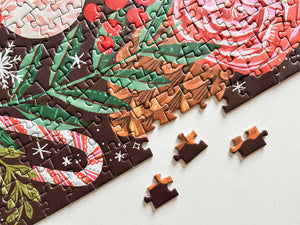 A close-up of a completed jigsaw puzzle showing a holiday floral design with red roses, green holly leaves, candy canes, and snowflakes on a dark brown background. Several loose puzzle pieces are scattered on a white surface.