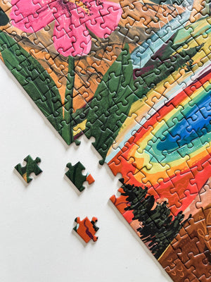 A partially completed jigsaw puzzle showing a colorful landscape with a rainbow, green trees, and pink flowers, with a few loose pieces on a white surface.