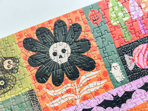 A close-up of a completed jigsaw puzzle featuring a colorful Halloween-themed patchwork design with a large black flower and white skull in the center, surrounded by orange, green, pink, purple, and black sections.