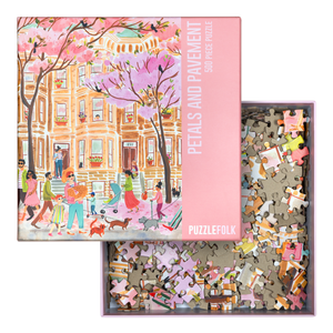 Puzzle box lid showing colorful illustration of a city street scene with pink cherry blossoms, pedestrians, and dogs on a pink background. Lid sits on an open box showing puzzle pieces. 