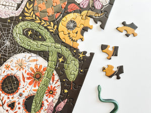 A partially assembled jigsaw puzzle featuring a Day of the Dead theme with a green snake, orange flowers, and a yellow skull on a dark background, shown on a white surface with a few loose pieces beside it.