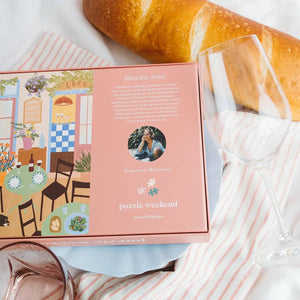 Puzzle box with a scenic cafe design, bread, and wine glasses on a tablecloth. Box shows details about the puzzle artist Elsa Eleni.