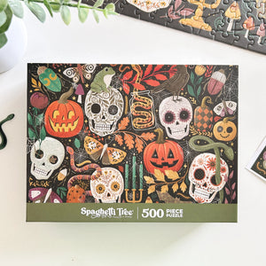 A 500-piece jigsaw puzzle box by Spaghetti Tree Jigsaw Puzzles, featuring a busy Halloween-themed design with sugar skulls, jack-o'-lanterns, candles, and snakes on a black background, displayed on a white surface.