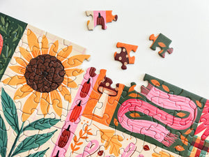 A colorful autumn-themed jigsaw puzzle partially assembled on a white surface, with several loose puzzle pieces scattered above it. The design features a large sunflower, pumpkins, leaves, and a pink scarf in warm orange, yellow, green, and pink tones.