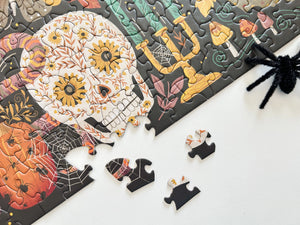A partially assembled jigsaw puzzle featuring a colorful sugar skull and Halloween-themed illustrations on a dark background, with a few loose puzzle pieces on a white surface and a black spider decoration in the corner.