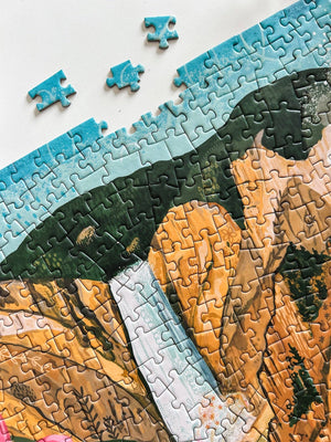 A close-up of a completed jigsaw puzzle showing a landscape scene with blue sky, green hills, and golden-brown cliffs, with a few loose puzzle pieces on a white surface.