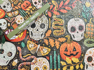 A 500-piece jigsaw puzzle box placed diagonally over a completed Halloween-themed puzzle featuring sugar skulls, pumpkins, leaves, spiders, and candles on a dark background.