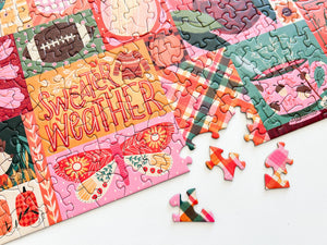 A colorful jigsaw puzzle with illustrated panels and the words 'SWEATER WEATHER' on an orange section, shown partially assembled on a white surface with a few loose puzzle pieces nearby.