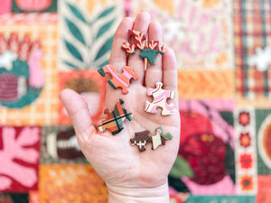 A hand holds several small, colorful, uniquely shaped wooden charms or pendants against a out of focus partial shot of the completed puzzle the pieces come from.