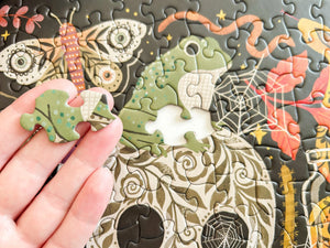 A close-up of a hand holding a small puzzle piece in front of an illustrated jigsaw puzzle featuring a green frog, butterflies, flowers, and spiderwebs on a dark background.