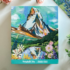Jigsaw puzzle box with colorful illustration of the iconic Matterhorn peak and flowers and lake in foreground on a white surface.