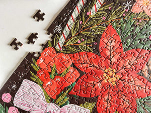 A close-up of a completed jigsaw puzzle showing a festive holiday design with large red poinsettias, green pine branches, pink bows, and dark brown background pieces, with a few loose puzzle pieces on a white surface.