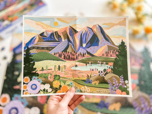 Hand holding a poster of Mountains & Valley puzzle artwork showing illustrated mountain and valley scene. 
