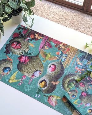 A colorful jigsaw puzzle featuring whimsical bird nests with flowers and small birds on a teal background, displayed on a white tabletop next to a matching puzzle box, with a potted plant and carpet visible.
