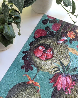Close up of finished portion of puzzle showing three birds inside a birdhouse made of grasses atop a blue-green patterned background. Puzzle is atop a white surface next to some plants.