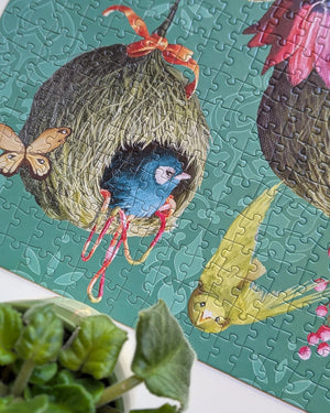 A close-up of a completed jigsaw puzzle featuring a blue bird in a brown nest with red accents, a yellow bird, and a butterfly on a teal background. A small green plant is visible in the lower left corner.