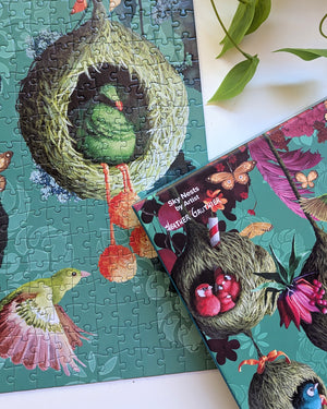 Partial view of puzzle box and completed puzzle showing whimsical bird houses made of grasses with birds and florals. The box shows puzzle details 'Sky Nests by artist Heather Gauthier in white text over image of the puzzle.  