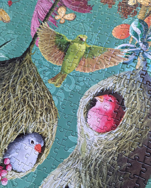 Partial view of completed puzzle with art of a green bird flying while a red and blue bird sit inside hanging bird houses made of grass. 