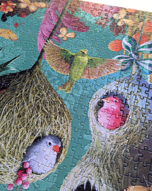 A close-up of a colorful jigsaw puzzle featuring birds in nests, with a green bird in flight and pink and gray birds in straw nests against a teal background.