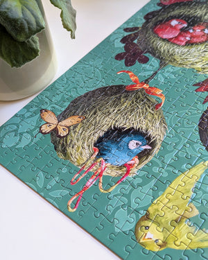 A completed jigsaw puzzle featuring a whimsical illustration of a blue bird inside a nest with red strings, placed on a white surface next to a white pot with green leaves.