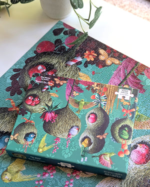 Puzzle box atop a partial view of the completed puzzle on white surface with view of part of a plant showing. Puzzle features painting of colorful birds flying around and resting in hanging bird houses. Bird houses are shown against a green-blue patterned background with feathers and flowers hanging amongst houses.
